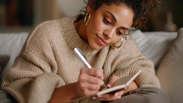 Young woman writing notes in a small book, journaling thoughts and ideas, focusing on self care, personal reflection, and cozy lifestyle at home for well being mindfulness