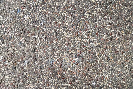 ​Top-down view of a textured exposed aggregate concrete surface with multicolored river pebbles and small stones embedded in gray cement, ideal for architectural backgrounds and paving textures.