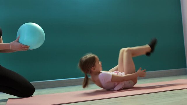 Instructor is holding stability ball while young girl performs supine flexibility stretch on yoga mat in fitness studio