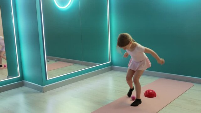 Young girl is practicing balance training on yoga mat using wobble cushion and massage ball in fitness studio with mirror, improving coordination and stability