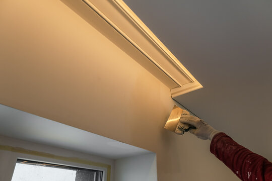 A close-up of a craftsman's hand in a white protective glove applying putty to a wall with a metal putty knife during an interior renovation.