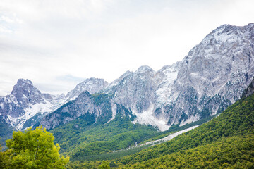Obraz premium Impressive rocky mountain peaks lightly covered with snow rise above a vibrant green forested valley, creating a powerful alpine landscape under a bright sky. The contrast between rugged cliffs