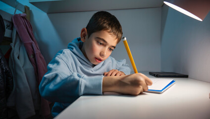 Young boy concentrating while writing in a notebook at his bedroom desk under a white lamp, during a quiet moment of studying and doing homework in the evening