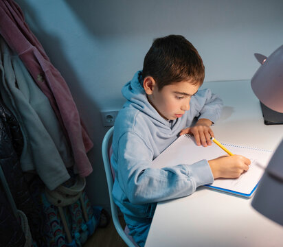 Young boy doing his homework alone at his desk in a cozy bedroom with warm lighting and focused expression during evening study time