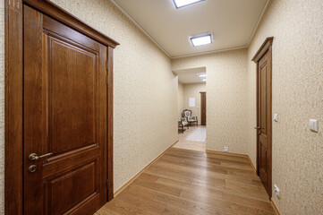 Fototapeta premium A long, light-toned hallway with wooden flooring, dark wooden doors, beige patterned wallpaper, recessed ceiling lights, leading to a seating area in the background.