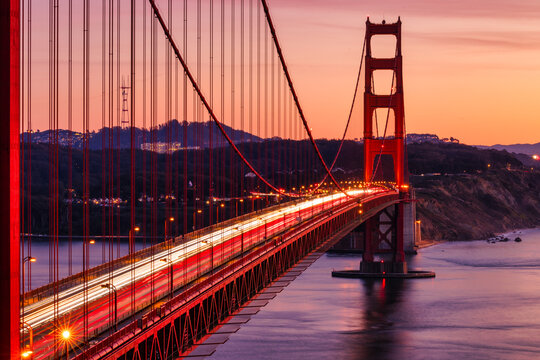 Long exposure view of the Golden Gate Bridge at dusk with flowing traffic light trails. Iconic red suspension bridge over calm bay water under a warm pastel evening sky