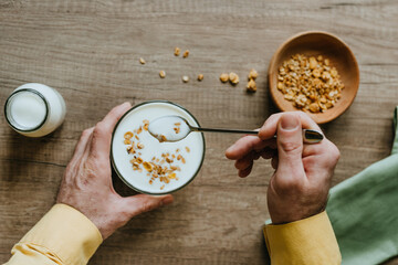 Top view of man adding granola to yogurt bowl on wooden background