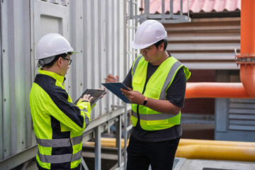 Two professional Asian technicians inspect an industrial cooling tower system using a digital tablet and clipboard. They perform maintenance checks on outdoor chiller units at a factory plant.