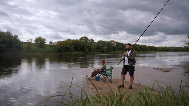 Happy young couple spending quality time together fishing rods from sandy river bank on cloudy day. Romantic outdoor activities and a shared hobby to connect with nature