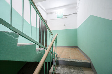 Narrow residential staircase with mint-green painted walls and steps, metal and wooden railings, tiled floor, dim lighting, dark doorway at the end. © evgeniykleymenov