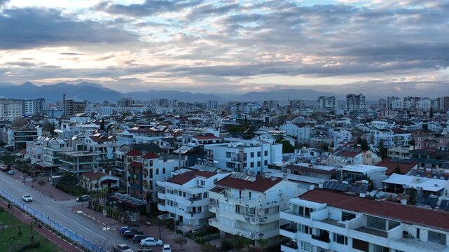 Aerial drone footage of Antalya&rsquo;s coastline in Turkey at sunset, showing modern seaside buildings rising above rocky cliffs along the Mediterranean Sea. Soft evening light colors the sky while the cit