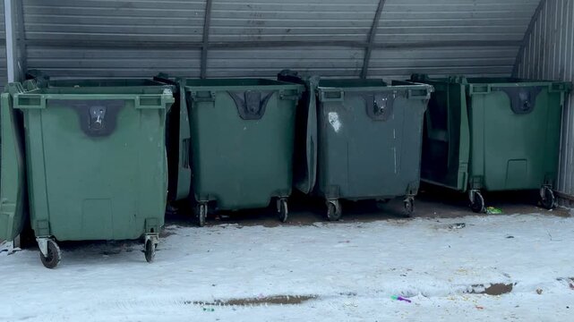 Dark green garbage bins in courtyard, lined under metal shelter on snow dusted concrete, empty wheels and closed lids, chipped paint and small stickers, quiet urban morning near apartment
