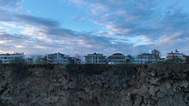 Aerial drone footage of Antalya&rsquo;s coastline in Turkey at sunset, showing modern seaside buildings rising above rocky cliffs along the Mediterranean Sea. Soft evening light colors the sky while the cit