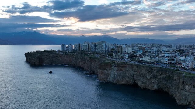 Aerial drone footage of Antalya&rsquo;s coastline in Turkey at sunset, showing modern seaside buildings rising above rocky cliffs along the Mediterranean Sea. Soft evening light colors the sky while the cit