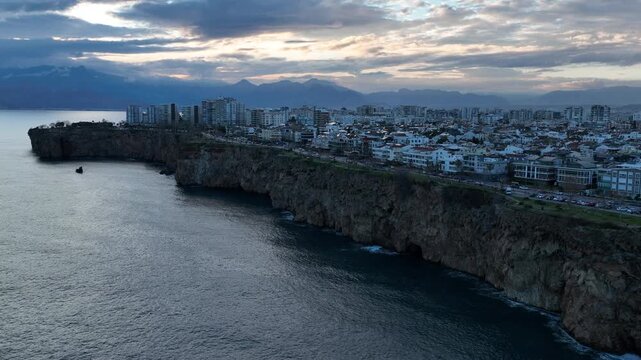 Aerial drone footage of Antalya&rsquo;s coastline in Turkey at sunset, showing modern seaside buildings rising above rocky cliffs along the Mediterranean Sea. Soft evening light colors the sky while the cit