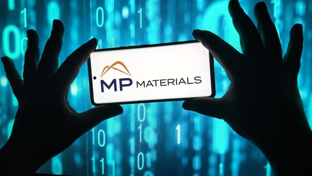 Hands hold smartphone displaying mp materials logo against binary code
