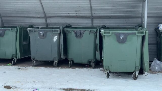 Dark green garbage bins in courtyard, lined under metal shelter on snow dusted concrete, empty wheels and closed lids, chipped paint and small stickers, quiet urban morning near apartment