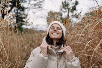 Obraz premium Woman in a warm beige coat and knit hat stands among tall grasses, smiling under a cloudy sky, capturing a cozy outdoor moment in late autumn or early winter soft breeze.