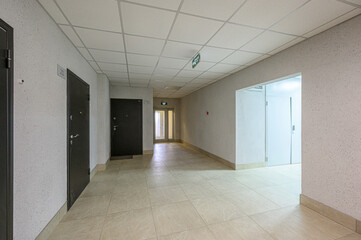 Fototapeta premium Long, light-toned corridor with tiled floor, textured walls, dark metal doors, white ceiling tiles, exit sign, clean, minimalist design, natural light at the end.