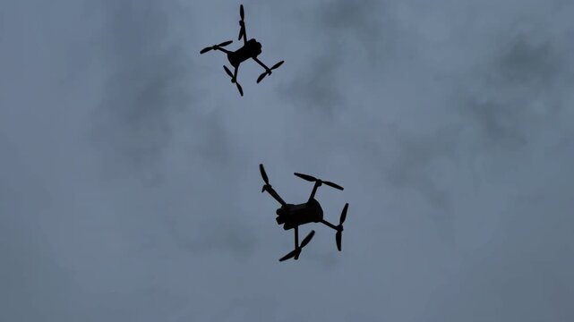 Two drones fly in a cloudy sky above an open area during daytime hours. Drones are seen flying in the cloudy sky