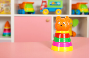 Fototapeta premium Colorful stacking toy shaped like a bear with rings in various colors placed on a pink table inside a kindergarten classroom with toy vehicles in the background