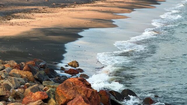 Waves wash over rocks on beach during the early morning sunlight. Waves gently roll onto the beach, meeting rocks as the sun rises over the horizon.
