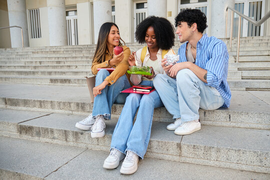 Diverse young students sitting on university stairs, eating healthy lunch and talking, enjoying a break