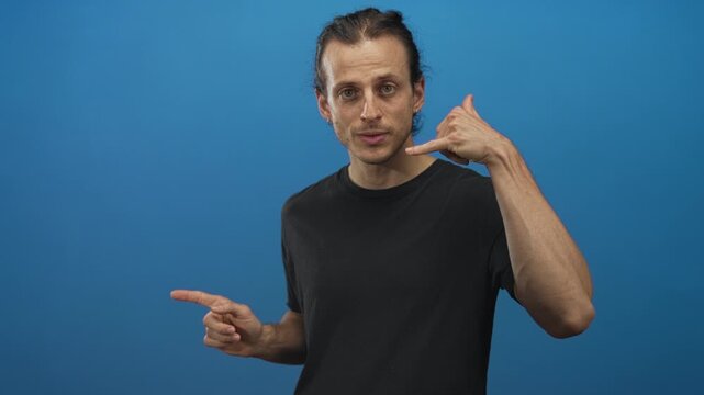 Man with long hair pointing finger and making phone gesture in studio with blue backdrop; playful invitation.