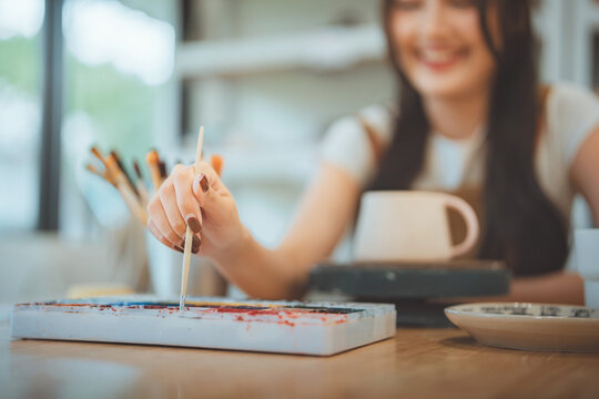 Close-up of a smiling Asian woman's hand using a paintbrush to paint on a ceramic mug in a creative workshop