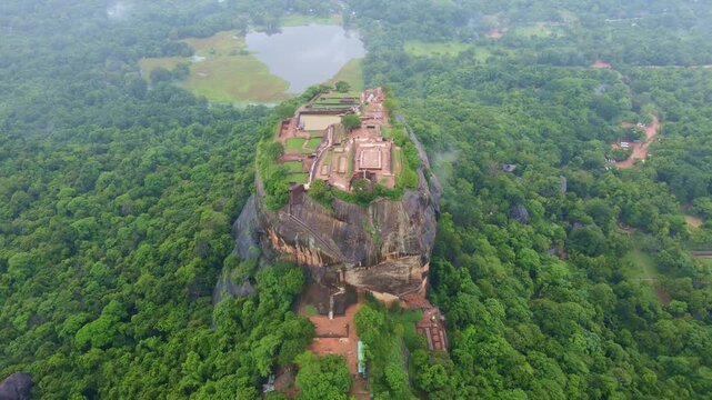 Early morning aerial drone footage of Sigiriya Rock Fortress after rain, Sri Lanka. Low clouds and mist drifting above lush tropical jungle, with wet surfaces reflecting soft morning light.Atmospheric