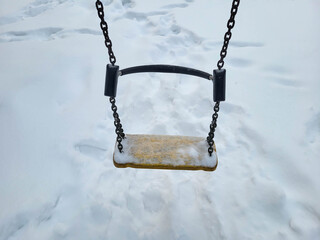 An empty swing seat in the snow on a playground hangs from metal chains on a cold winter day