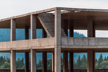 Partially constructed reinforced concrete building, shown as a raw structural frame without walls or finishes. In the background, forested hills and mountain silhouette