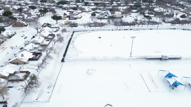 Football track field near Mockingbird Elementary complex, residential homes appear uniformly snow-covered. Light poles, fences, blue playground canopies Winter Storm Fern white icy, Coppell, TX