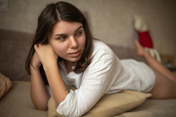 Fototapeta premium Young woman with long dark hair resting on a couch, wearing a white shirt, with soft pillows and a neutral background creating a cozy indoor atmosphere