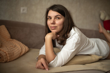 Fototapeta premium Young woman with long dark hair resting on a couch, wearing a white shirt, with soft pillows and a neutral background creating a cozy indoor atmosphere