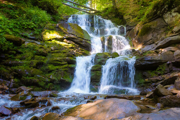 Obraz premium waterfall in the forest on a summer morning. rocks with moss in dappled light. refreshing nature scenery