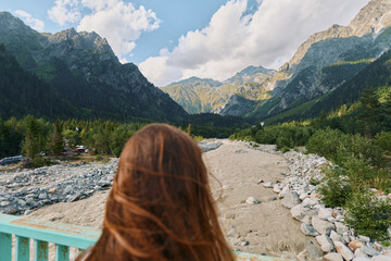 A person with long brown hair stands on a bridge overlooking a rocky river valley framed by towering mountains. They gaze outward, enjoying tranquil natural scenery and peaceful solitude. © SHOTPRIME STUDIO