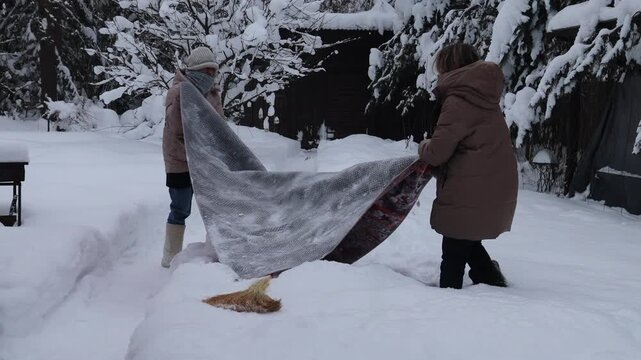 Close-up footage of a mother and daughter cleaning and beating a rug with a broom outdoors on fresh snow. Winter housekeeping and traditional carpet cleaning, with snow flying in the air and focus on 