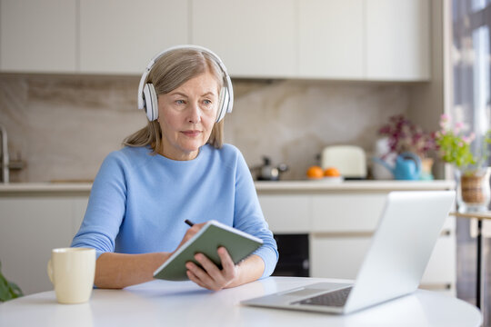 Senior woman at home with headphones, focused on laptop, taking notes during an online course or video call engaged in remote learning, work, or self-improvement activities