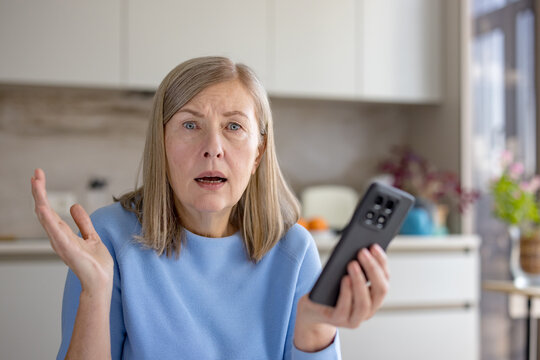 Elderly woman with a shocked expression, holding a smartphone and gesturing with her hand, experiencing frustration and tech difficulties in a modern kitchen setting