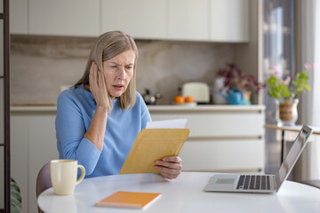 Senior woman sitting at her kitchen table with a laptop, reading a distressing bill in an opened...