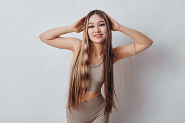 Fototapeta premium Young woman with long braided hair posing with hands on head wearing beige tank top and leggings against white background. Beauty, confidence and fashion concept in studio.