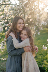 Fototapeta premium A young Caucasian woman with long brown hair embraces a young girl with curly brown hair in a blooming garden. Soft sunlight filters through the flowers.