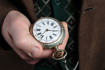 Close-up of a man's hand holding a classic pocket watch with Roman numerals against the background...