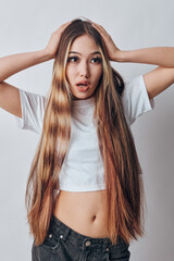 Young woman with long hair and casual clothes posing with hands on head and surprised facial expression in studio on light background. Beauty, emotion and fashion portrait.