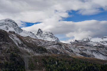 Majestic snow-capped mountain peaks and rocky slopes under a dramatic cloudy blue sky in the Alps, featuring thin hiking trails and pine forests.