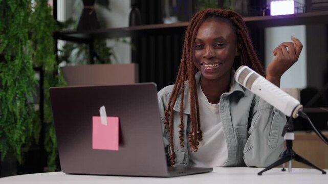 Portrait African female adult presenting podcast from modern office with laptop and microphone, online video streaming broadcast or videoblog.