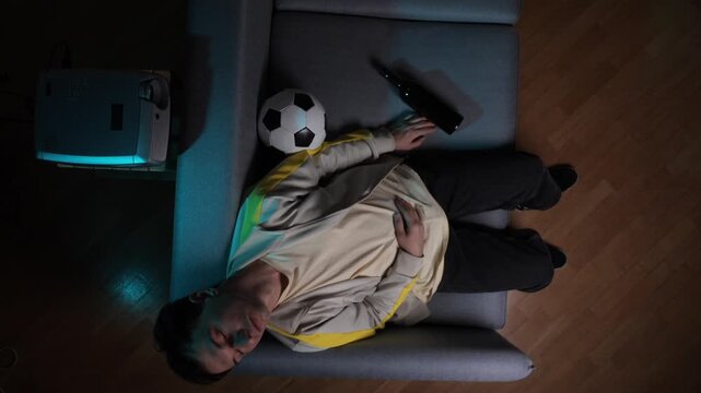 Exhausted male football fan asleep on grey sofa at home, soccer ball and bottle nearby, projector casting blue light in dark living room after a late night match
