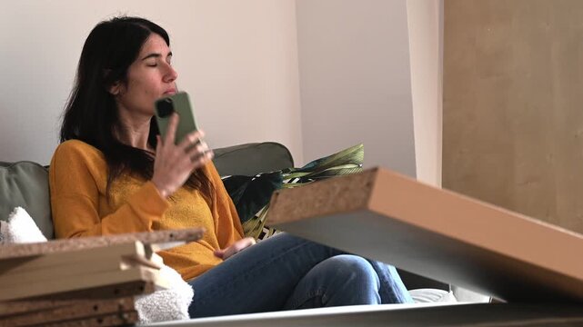 Young woman sitting on a sofa using her smartphone to call a furniture removal service, surrounded by old furniture at home
