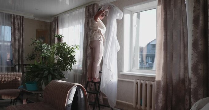 A housewife stands on a stepladder in the living room, removing white tulle curtains from the curtain rod for washing and cleaning. The woman is doing a general spring cleaning of the house.
